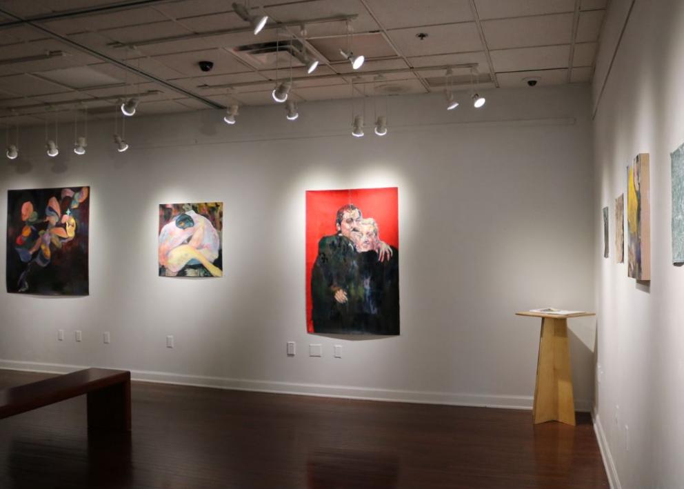 Multiple paintings on display throughout the gallery. From left to right: colorful swirls with a black background, abstract two naked bodies hugging one another on the ground, two humans side hugging one another starting to morph into one with black torsos and detailed heads. 