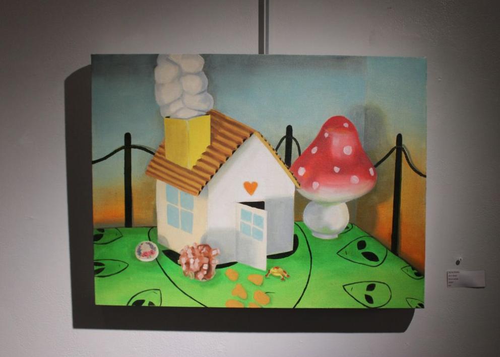 A white small home on top of a green alien tablecloth with a frog exiting from the front door. Around the house there are stones, a painted rock, and a mushroom the size of the house. 