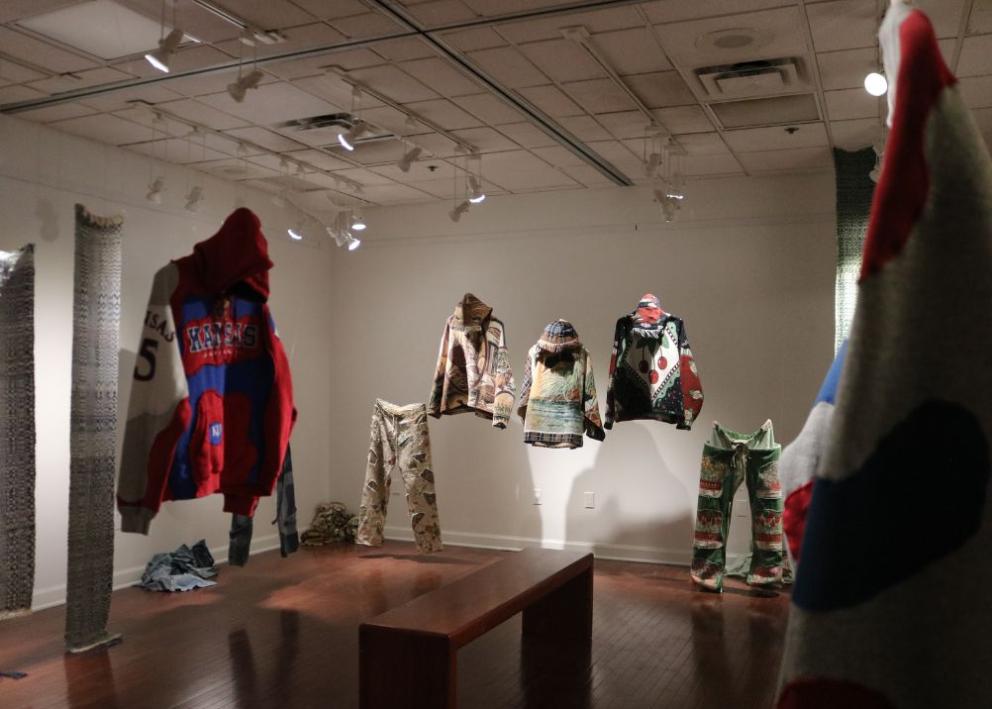 Multiple articles of patchwork clothing spaced around the gallery on display. Includes a Univeristy of Kansas Sweatshirt, sweatpants with flower pots, cherry and flower sweatshirt, beachfront lake and flannel sweatshirt, and a wildlife animal sweatshirt. 
