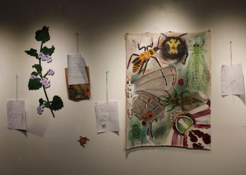 From left to right: a felt made vine of white flowers and dark green leaves, a paper made beetle, and a large painted canvas of various insects including a bee, butterfly, 
